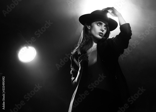Fototapeta Beautiful braless actress girl, wearing an unbuttoned blazer and a hat, poses and dances in the warm light rays of scenic illuminators in the theatrical fog