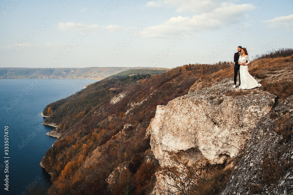 Obraz premium Beautiful bride on a mountains and river background. Cute girl in white dress. Mountains landscape and rivers in the sunset