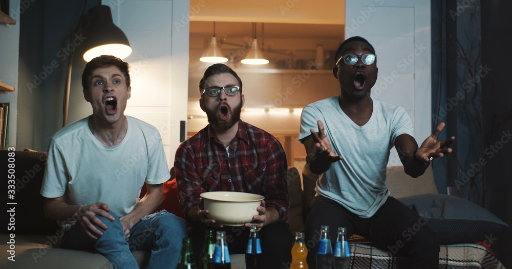 © vadim_key - Happy multiethnic young men watch sports game, cheer together at home with snacks using big projector screen slow motion