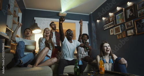 Excited young happy multiethnic friends get emotional and shout watching sports on TV together at home slow motion.
