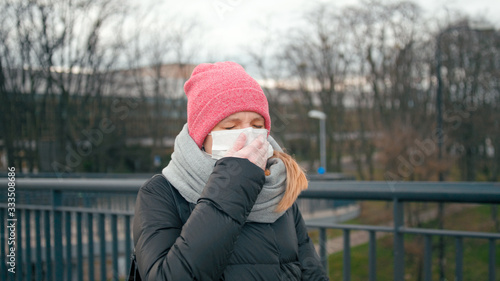 COVID-19 Coronavirus Pandemic Outbreak: Casual Young Sick Woman in Surgical Face Mask and Medical Gloves Sneezes in Street and Coughs covering mouth