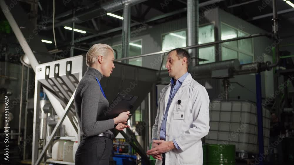 Side view of female manager or journalist with clipboard and male engineer in white coat talking in factory production department Side view of female manager or journalist with clipboard and male engineer in white coat talking in factory production department