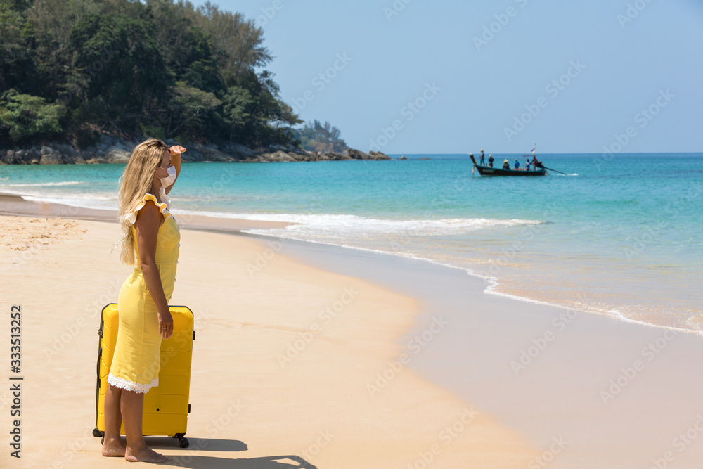 A woman in a protective surgical mask on her face, walking along the beach with a yellow suitcase. Chinese coronavirus disease COVID-19 is a dangerous virus. Pandemic.