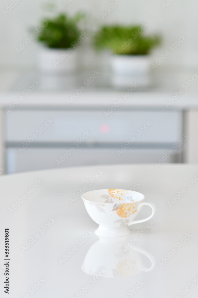 Working alone at the office, coffee cup on a white table over blurred clean kitchen with no one and no clients in a cafe, a reflection of the floral cup is reflecting from the table Working alone at the office, coffee cup on a white table over blurred clean kitchen with no one and no clients in a cafe, a reflection of the floral cup is reflecting from the table