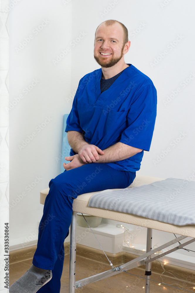 Fototapeta premium Portrait of a young male doctor. Man in blue uniform of doctor sits on massage table in massage room