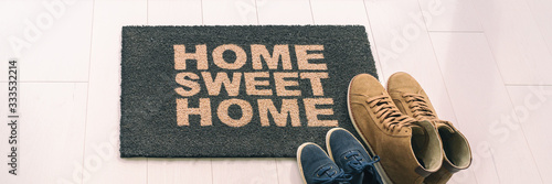 Home sweet home entrance door mat at condo floor with couples pairs of shoes moving in together. women's shoes and man's boots on floor, new apartment. Panoramic banner.