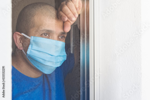 Sick man with face mask looking out the window being quarantined at home. He is sad that he cannot get out due to the Covid-19 coronavirus pandemic.Space for text.