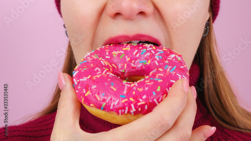 Beautiful adult female in sweater and hat biting delicious donut against pink background. Close up