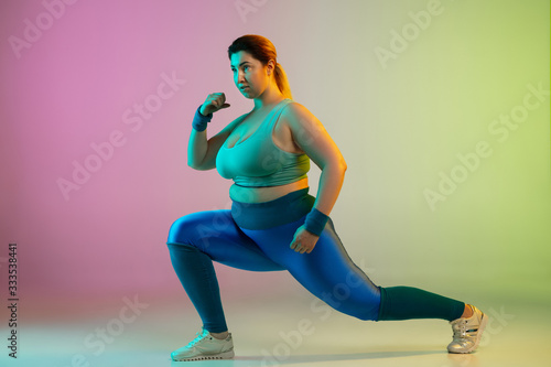Young caucasian plus size female model's training on gradient purple green background in neon. Doing stretching workout exercises. Concept of sport, healthy lifestyle, body positive, equality.