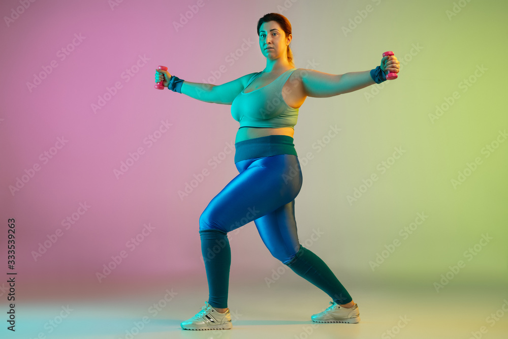Young caucasian plus size female model's training on gradient purple green background in neon light. Doing workout exercises with weights. Concept of sport, healthy lifestyle, body positive, equality.