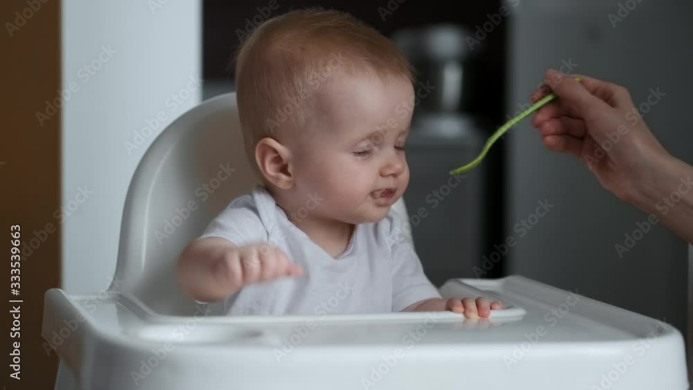 Cute little baby feeding with a spoon at the table, the baby smiles at the camera and then cries and gets cranky infant eating meal foods. 4k 50 fps Cute little baby feeding with a spoon at the table, the baby smiles at the camera and then cries and gets cranky infant eating meal foods. 4k 50 fps