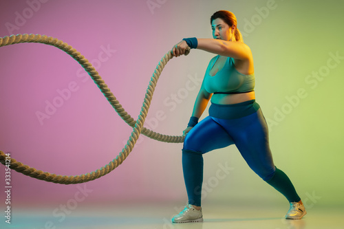 Young caucasian plus size female model's training on gradient purple green background in neon light. Doing workout exercises with ropes. Concept of sport, healthy lifestyle, body positive, equality. © master1305
