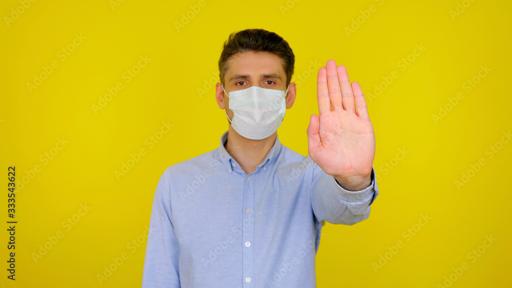 Stop virus concept: coronavirus, flu. Man in protective medical mask looks at the camera. The guy in the blue shirt shows stop on a yellow background Stop virus concept: coronavirus, flu. Man in protective medical mask looks at the camera. The guy in the blue shirt shows stop on a yellow background
