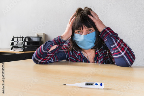 Sick woman with coronavirus disease and blue protective mask on face. Covid-19 pandemic outbreak. High fever on medical thermometer on table. Headache and panic fear in quarantine at home office work. photo