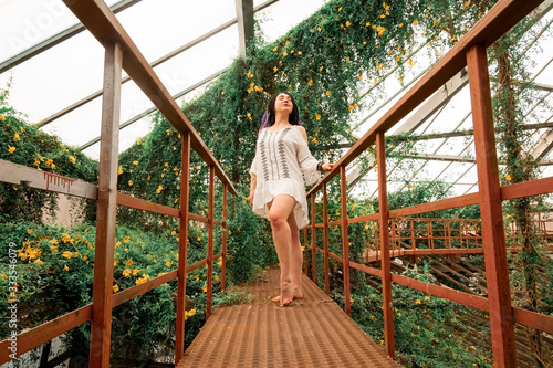 a lonely beautiful girl with multi-colored dreadlocks in a white dress stands on a red stairs in an arch of flowers. Greenhouse with exotic tropical plants in the botanical garden in Ukraine, Dnipro.
