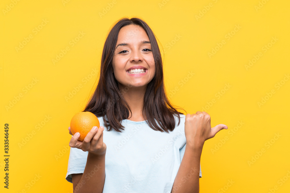 Young brunette woman holding an orange pointing to the side to present a product