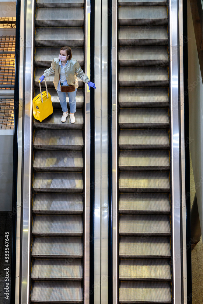 Obraz premium Woman with yellow luggage stands on escalator at almost empty airport terminal due to coronavirus pandemic/Covid-19 outbreak travel restrictions. Flight cancellation. Quarantine measures. Top view. 