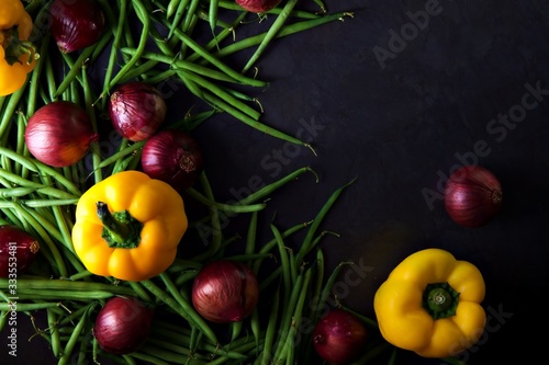 Green beans and red onions with yellow peppers scattered across a dark grey background. 