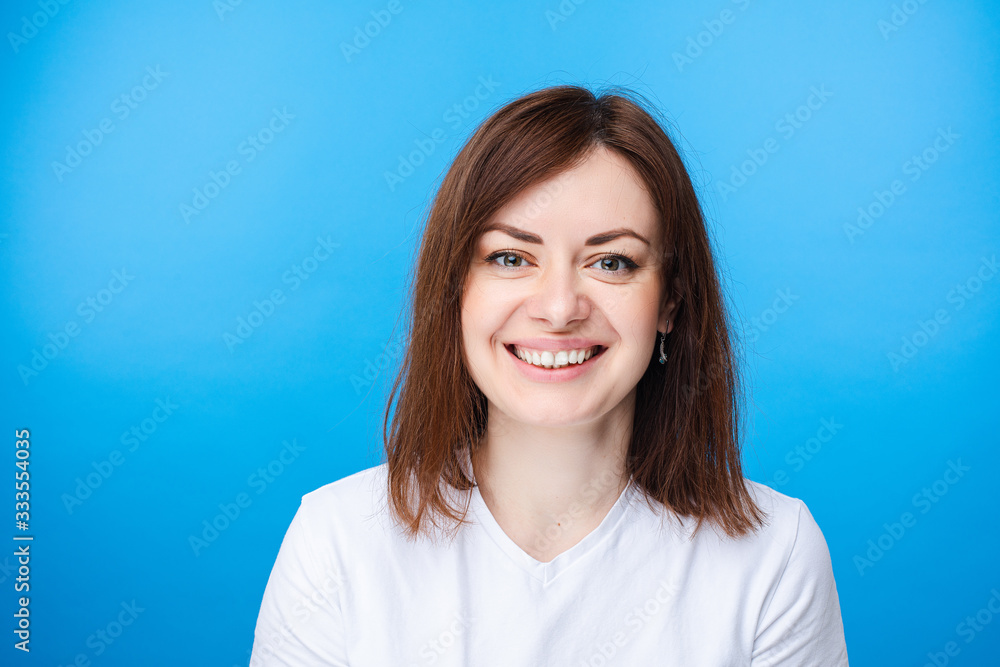 Stock studio portrait of a pretty happy young adult woman with middle-length dark hair in white t-shirt smiling at camera with toothy smile. Isolate on light blue background. Stock studio portrait of a pretty happy young adult woman with middle-length dark hair in white t-shirt smiling at camera with toothy smile. Isolate on light blue background.