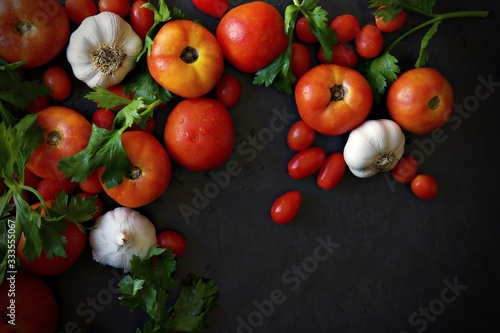 Large and cherry tomatoes with white onions, whole garlic and large leaf Italian parsley against a dark grey background. Landscape flat lay with space for text.