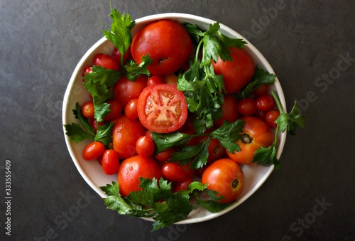 White bowl of large and cherry tomatoes with large leaf Italian parsley against a dark grey background. 