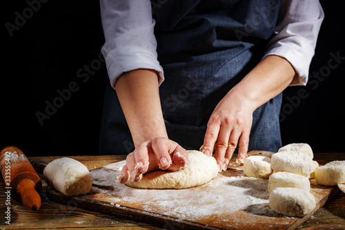 Female hands knead the dough on a wooden antique table on a dark background, close-up, shallow depth of field, beautiful directional lighting. Concept of home baking and comfort.