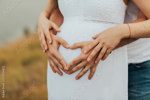 Cropped image of beautiful pregnant woman and her handsome husband hugging the tummy. Couple making a heart shape on the pregnant belly with their hands. Concept of pregnancy, expecting a baby, love. © eduard