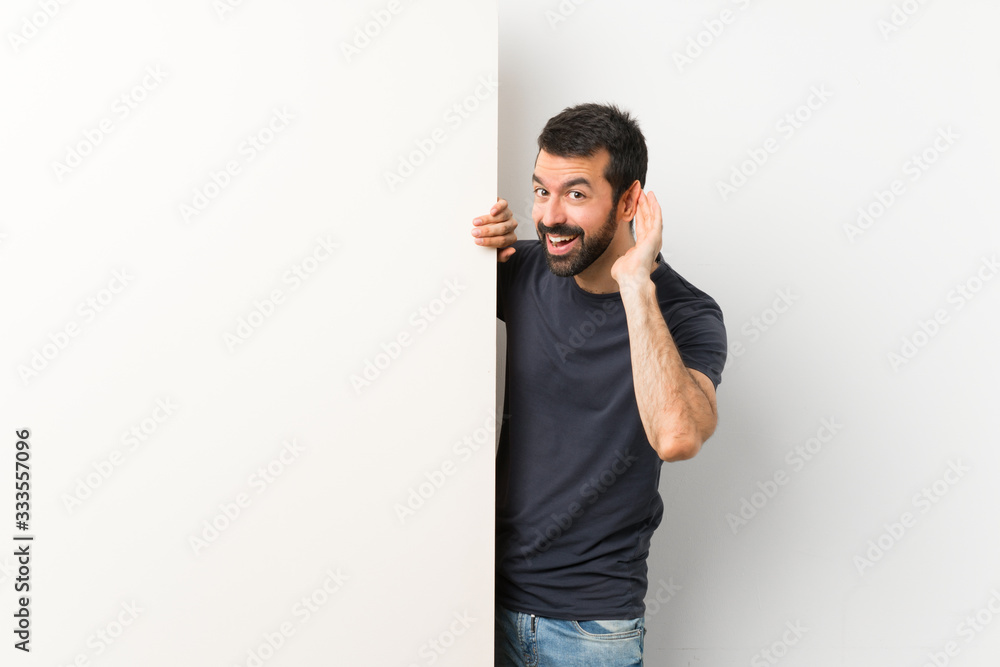 Young handsome man with beard holding a big empty placard listening to something by putting hand on the ear Young handsome man with beard holding a big empty placard listening to something by putting hand on the ear