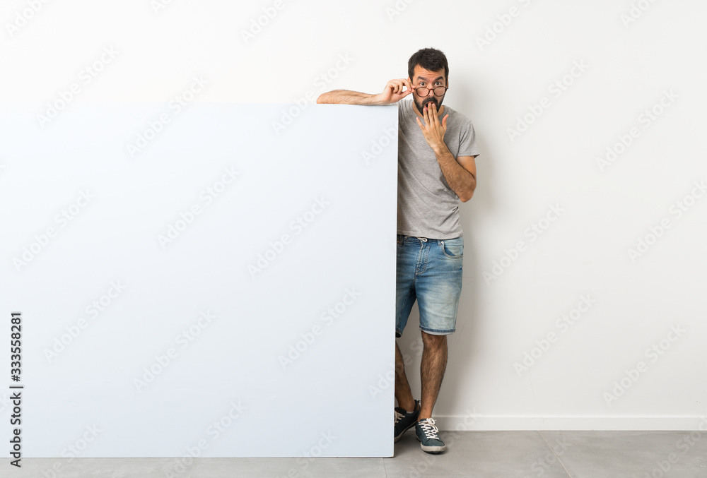 Young handsome man with beard holding a big blue empty placard with glasses and surprised Young handsome man with beard holding a big blue empty placard with glasses and surprised