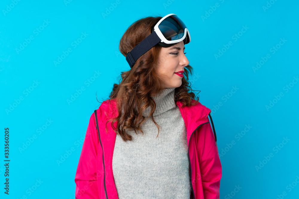 Skier woman with snowboarding glasses over isolated blue wall looking to the side Skier woman with snowboarding glasses over isolated blue wall looking to the side