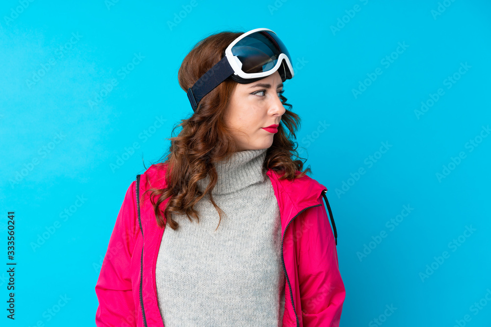 Skier woman with snowboarding glasses over isolated blue wall looking side Skier woman with snowboarding glasses over isolated blue wall looking side