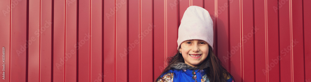 Happy girl kid with smartphone in hands on a red background in the street. Dressed in a blue jacket and smiling. Happy girl kid with smartphone in hands on a red background in the street. Dressed in a blue jacket and smiling.