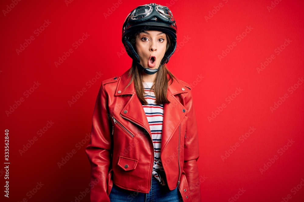 Young beautiful brunette motocyclist woman wearing motorcycle helmet and red jacket afraid and shocked with surprise expression, fear and excited face. Young beautiful brunette motocyclist woman wearing motorcycle helmet and red jacket afraid and shocked with surprise expression, fear and excited face.