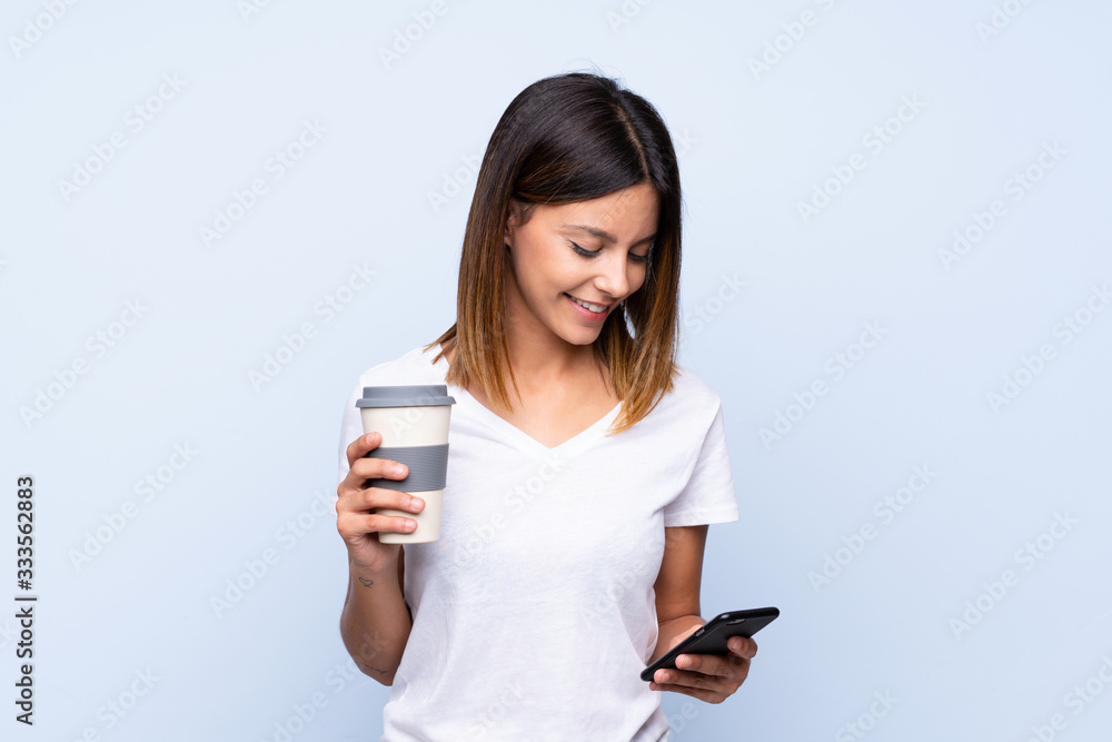 Young woman over isolated blue background holding coffee to take away and a mobile Young woman over isolated blue background holding coffee to take away and a mobile