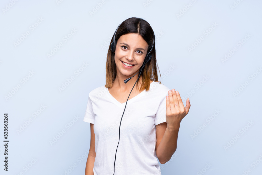 Young woman over isolated blue background working with headset doing coming gesture Young woman over isolated blue background working with headset doing coming gesture