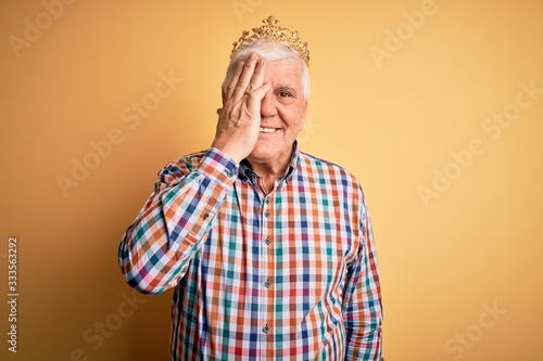 Senior handsome hoary man wearing golden crown of king over isolated yellow background covering one eye with hand, confident smile on face and surprise emotion.
