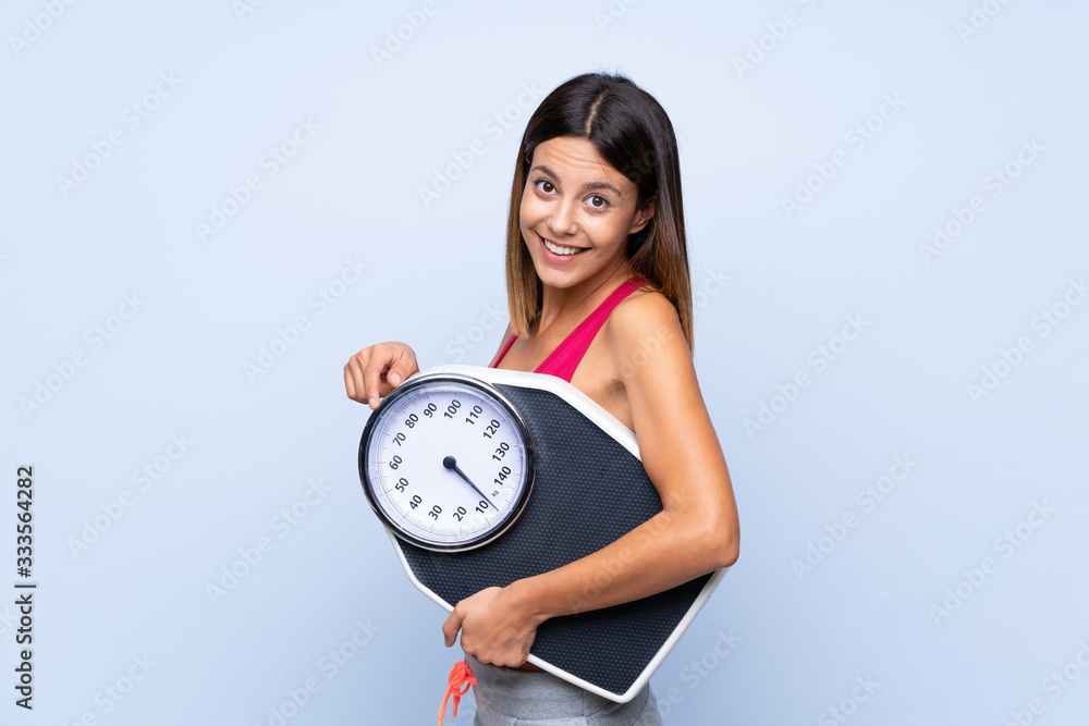 Girl with weighing machine over isolated blue background holding a weighing machine and pointing it Girl with weighing machine over isolated blue background holding a weighing machine and pointing it