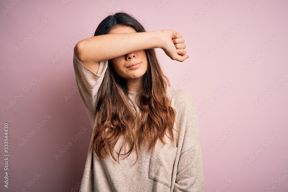 Fototapeta premium Young beautiful brunette woman wearing casual sweater standing over pink background covering eyes with arm, looking serious and sad. Sightless, hiding and rejection concept