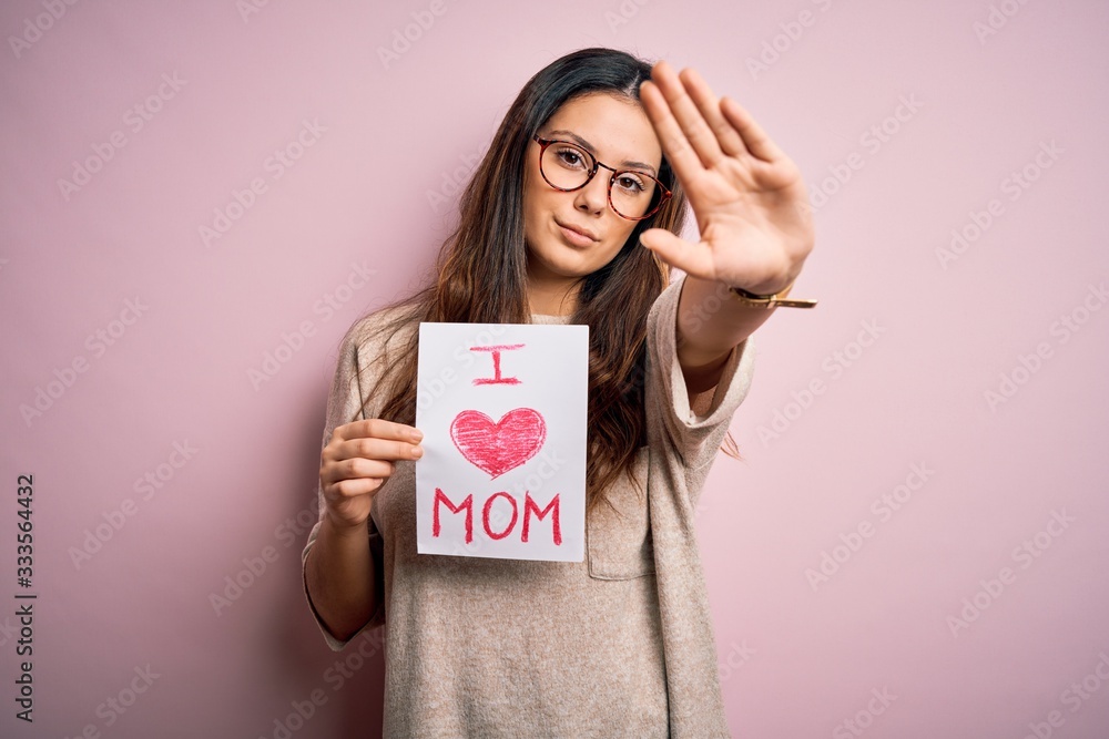 Young beautiful woman holding paper with love mom message celebrating mothers day with open hand doing stop sign with serious and confident expression, defense gesture Young beautiful woman holding paper with love mom message celebrating mothers day with open hand doing stop sign with serious and confident expression, defense gesture