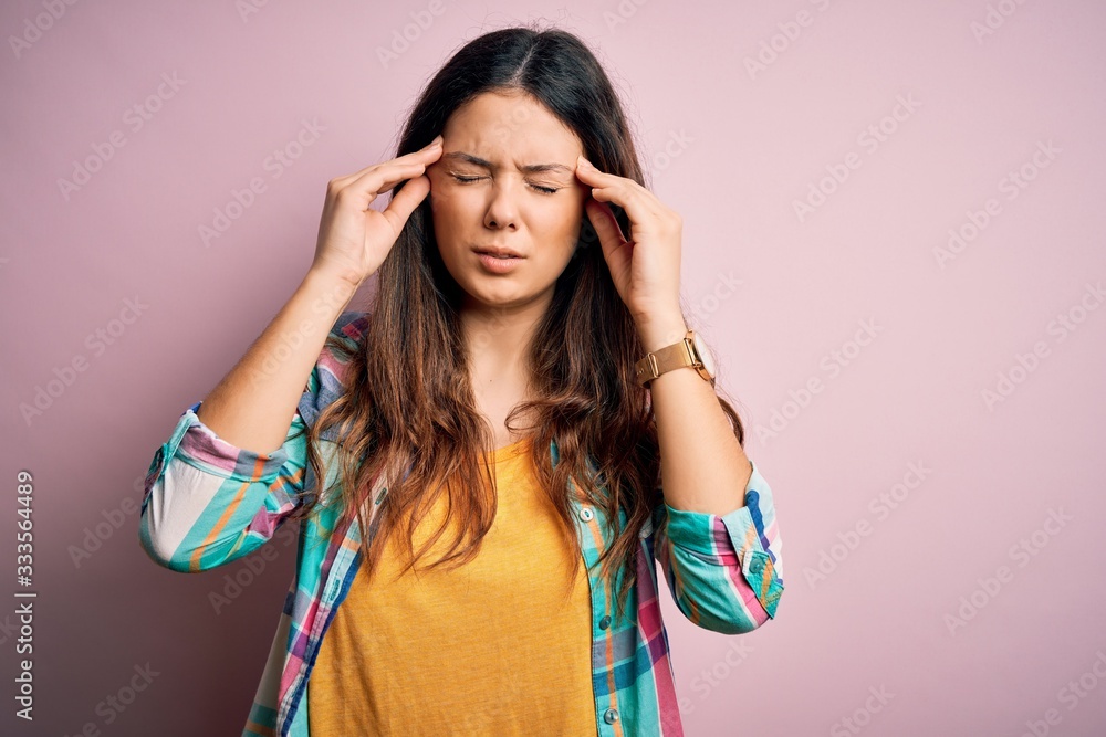Young beautiful brunette woman wearing casual colorful shirt standing over pink background with hand on head for pain in head because stress. Suffering migraine. Young beautiful brunette woman wearing casual colorful shirt standing over pink background with hand on head for pain in head because stress. Suffering migraine.