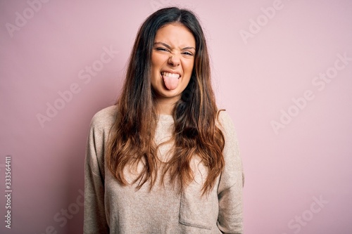 Young beautiful brunette woman wearing casual sweater standing over pink background sticking tongue out happy with funny expression. Emotion concept.