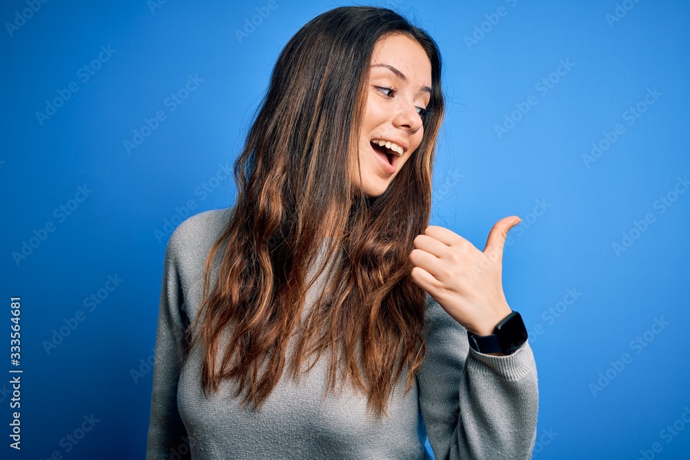 Fototapeta premium Young beautiful brunette woman wearing casual sweater standing over blue background smiling with happy face looking and pointing to the side with thumb up.