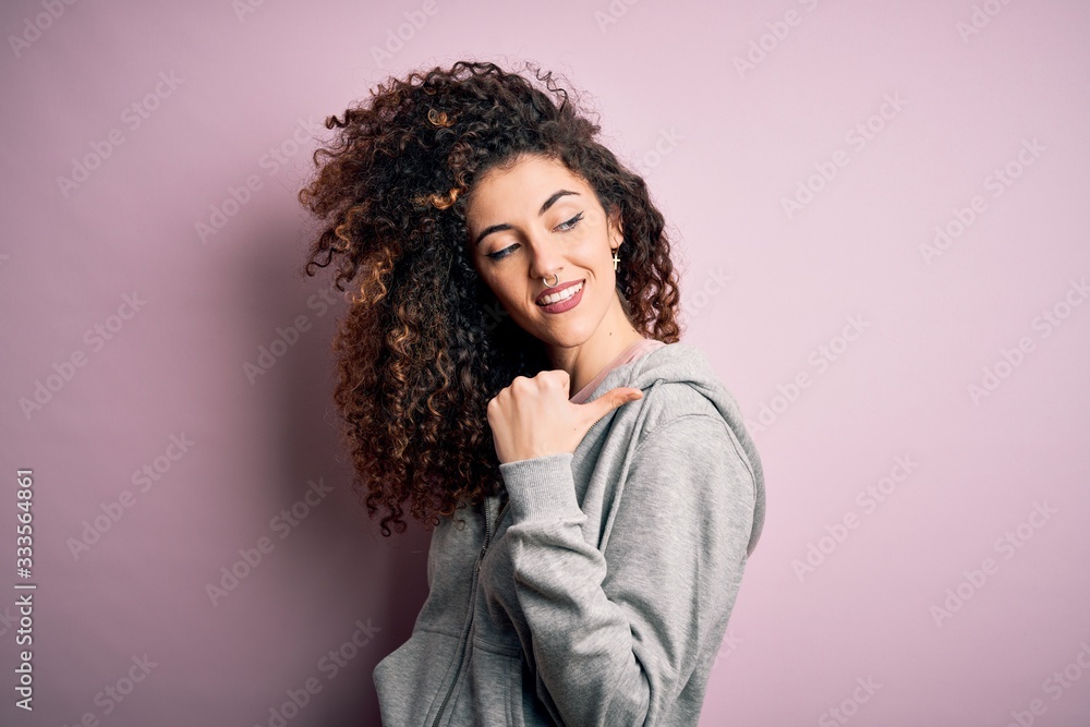 Young beautiful brunette sportswoman with curly hair and piercing wearing sportswear smiling with happy face looking and pointing to the side with thumb up. Young beautiful brunette sportswoman with curly hair and piercing wearing sportswear smiling with happy face looking and pointing to the side with thumb up.