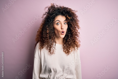 Beautiful woman with curly hair and piercing wearing casual sweater over pink background afraid and shocked with surprise expression, fear and excited face.