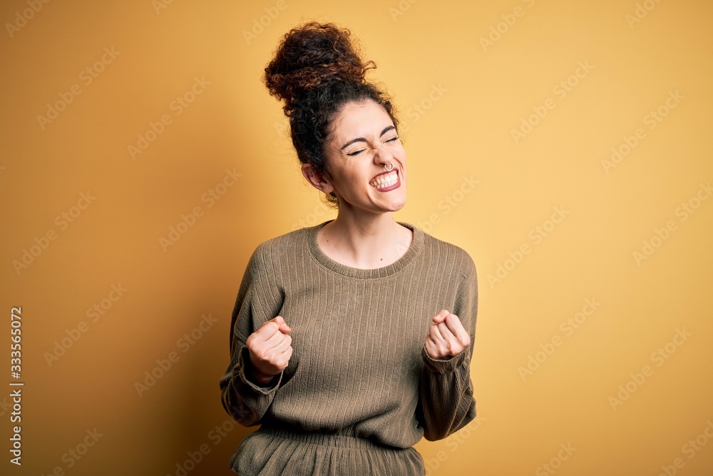 Young beautiful brunette woman with curly hair and piercing wearing casual dress excited for success with arms raised and eyes closed celebrating victory smiling. Winner concept. Young beautiful brunette woman with curly hair and piercing wearing casual dress excited for success with arms raised and eyes closed celebrating victory smiling. Winner concept.