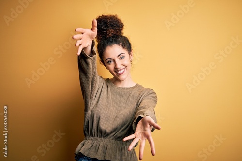 Young beautiful brunette woman with curly hair and piercing wearing casual dress looking at the camera smiling with open arms for hug. Cheerful expression embracing happiness. © Krakenimages.com