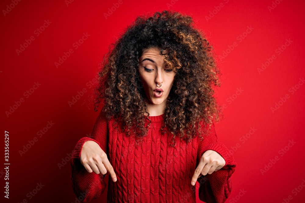 Young beautiful woman with curly hair and piercing wearing casual red sweater Pointing down with fingers showing advertisement, surprised face and open mouth Young beautiful woman with curly hair and piercing wearing casual red sweater Pointing down with fingers showing advertisement, surprised face and open mouth