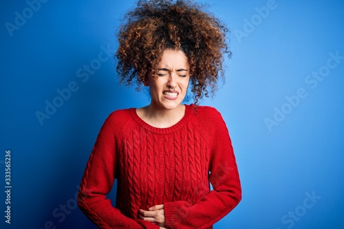 Young beautiful woman with curly hair and piercing wearing casual red sweater with hand on stomach because nausea, painful disease feeling unwell. Ache concept.