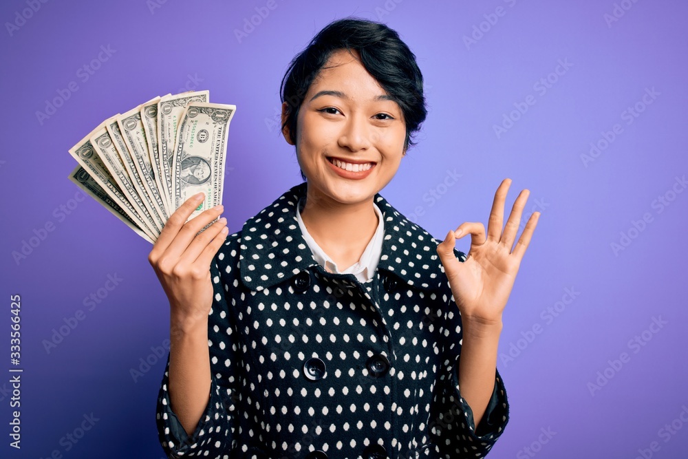 Young beautiful asian girl holding bunch of dollars banknotes over purple background doing ok sign with fingers, excellent symbol Young beautiful asian girl holding bunch of dollars banknotes over purple background doing ok sign with fingers, excellent symbol