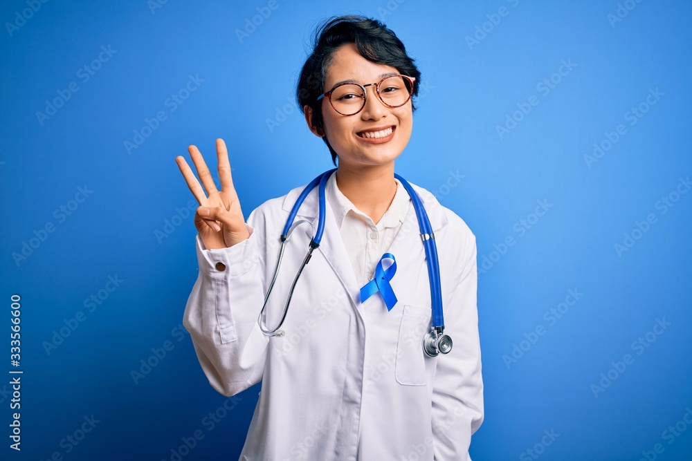 Young beautiful asian doctor girl wearing stethoscope and coat with blue cancer ribbon showing and pointing up with fingers number three while smiling confident and happy. Young beautiful asian doctor girl wearing stethoscope and coat with blue cancer ribbon showing and pointing up with fingers number three while smiling confident and happy.